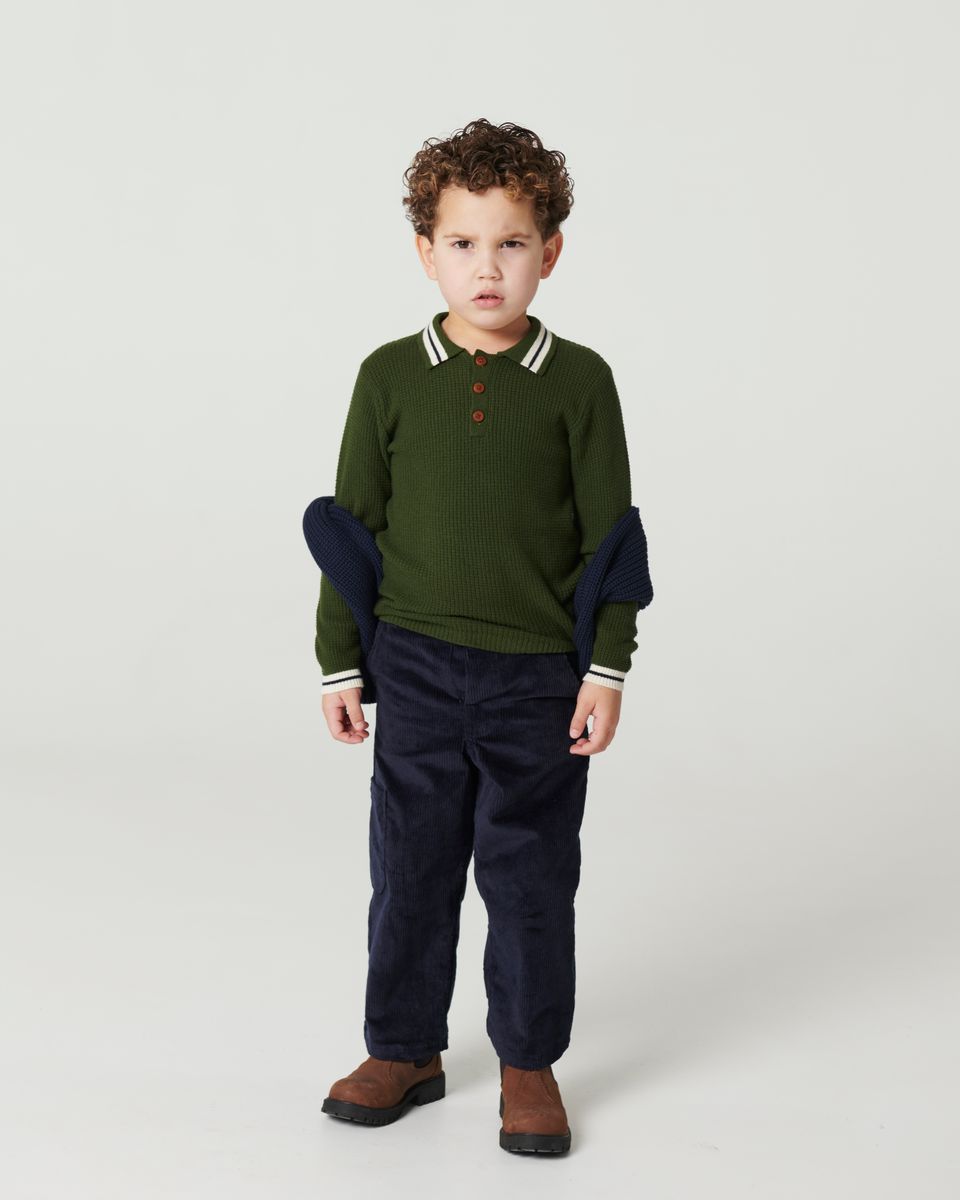 Young boy with curly hair wears Copenhagen Colors Boys’ Knitted Waffle Polo Long-Sleeved Polo, dark pants, and brown shoes, standing with a serious expression.