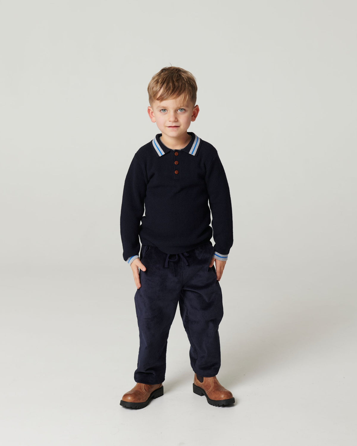 Young child on a plain background wears the Copenhagen Colors Boys’ Knitted Waffle Polo Long-Sleeved Polo, dark pants, and brown shoes, standing with hands by their sides.