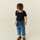 A young boy looks down, wearing a navy polo, Copenhagen Colors Baby Boys’ and Boys’ Organic Junior Jeans - Regular Fit, a jacket at his waist, and brown shoes on a beige backdrop.