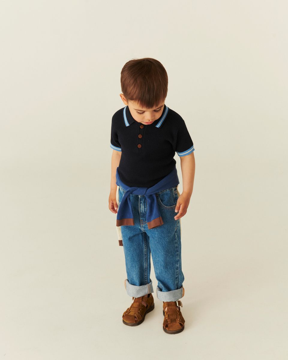A young boy looks down, wearing a navy polo, Copenhagen Colors Baby Boys’ and Boys’ Organic Junior Jeans - Regular Fit, a jacket at his waist, and brown shoes on a beige backdrop.