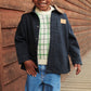 A smiling child in a navy jacket, plaid sweater, and Copenhagen Colors Baby Boys’ and Boys’ Organic Junior Jeans - Regular Fit stands by a wooden walkway.