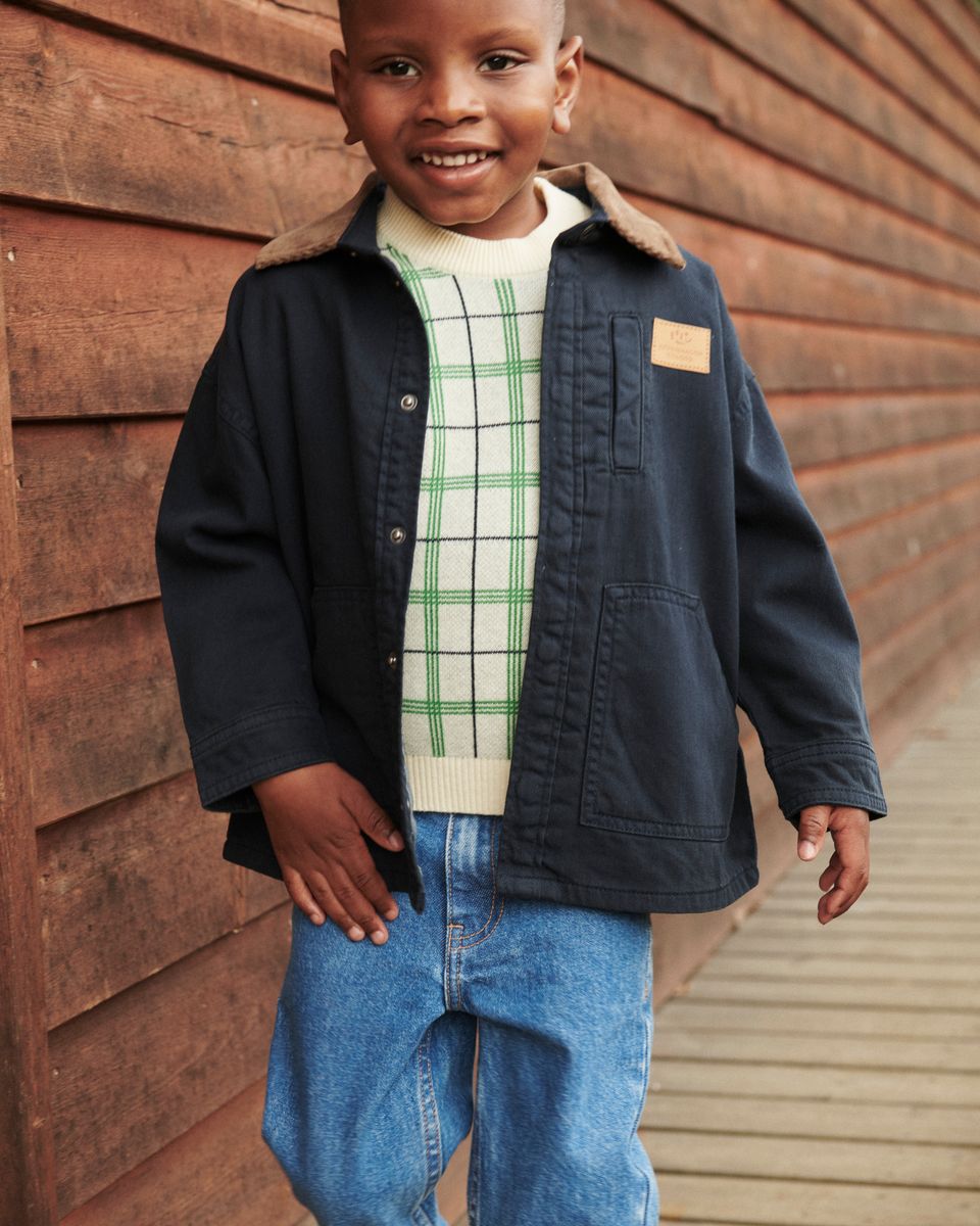 A smiling child in a navy jacket, plaid sweater, and Copenhagen Colors Baby Boys’ and Boys’ Organic Junior Jeans - Regular Fit stands by a wooden walkway.