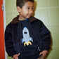 A young boy wearing the Copenhagen Colors Baby Boys’ and Boys’ Rocket Sweater stands by a green tiled wall, hands in pockets, looking to the side.