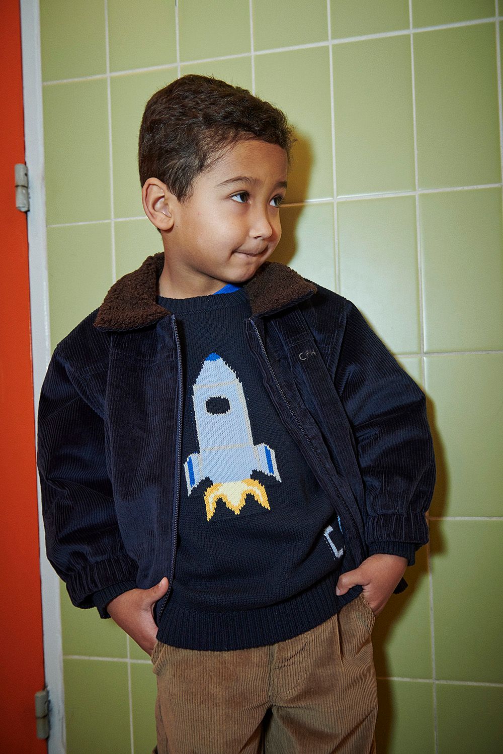 A young boy wearing the Copenhagen Colors Baby Boys’ and Boys’ Rocket Sweater stands by a green tiled wall, hands in pockets, looking to the side.