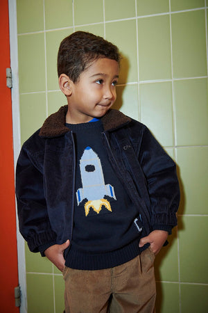 A young boy wearing the Copenhagen Colors Baby Boys’ and Boys’ Rocket Sweater stands by a green tiled wall, hands in pockets, looking to the side.