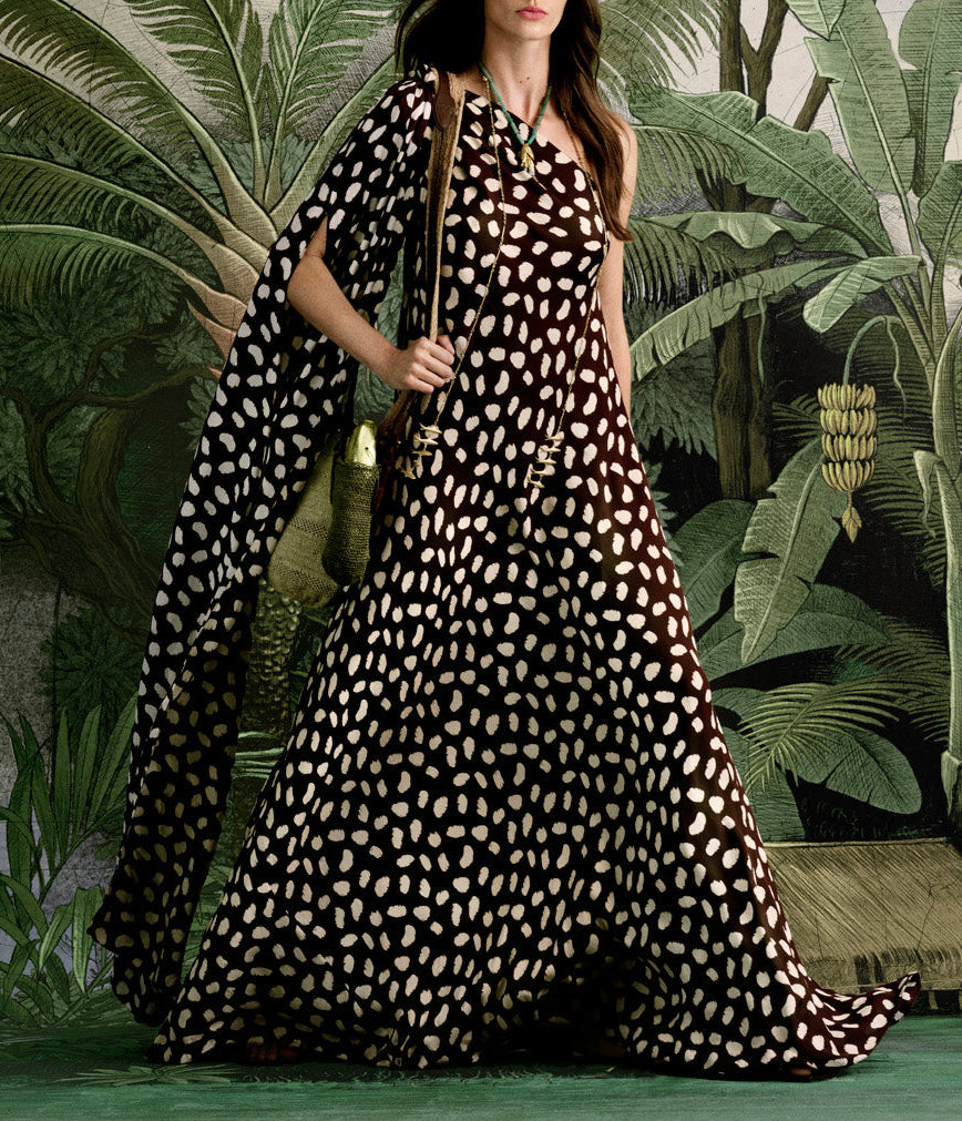 A woman stands before lush green tropical leaves wearing the Johanna Ortiz Silk Elegant Freedom Maxi Dress, a one-shoulder black and white polka dot design.