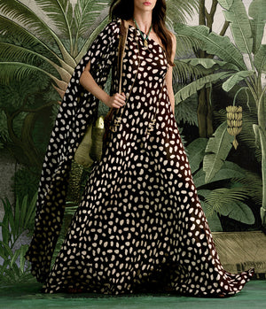 A woman stands before lush green tropical leaves wearing the Johanna Ortiz Silk Elegant Freedom Maxi Dress, a one-shoulder black and white polka dot design.
