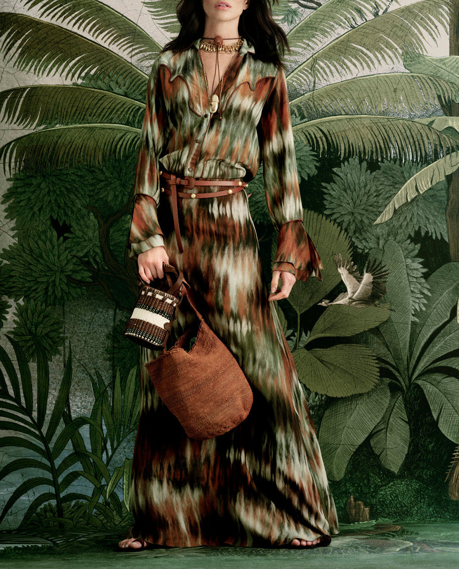 A woman in the Johanna Ortiz Cabalgando Shirt stands before tropical plants, holding two bags and wearing layered necklaces—perfect spring style with classic collar details.