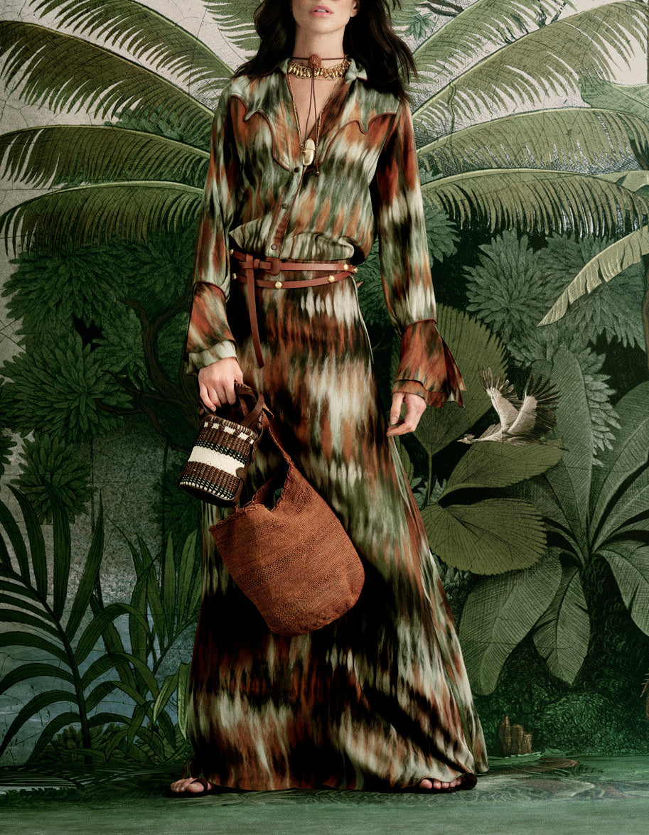 A woman stands before lush tropical plants, wearing the Johanna Ortiz New Intermingled Ideas Maxi Skirt in earth tones, paired with layered necklaces and holding two woven bags.