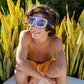 A teen boy smiles as he sits outdoors in front of tall green plants, wearing Bling2O Kids' Prismatic Goggles with UV protection and yellow swim trunks, his arms resting comfortably on his knees.