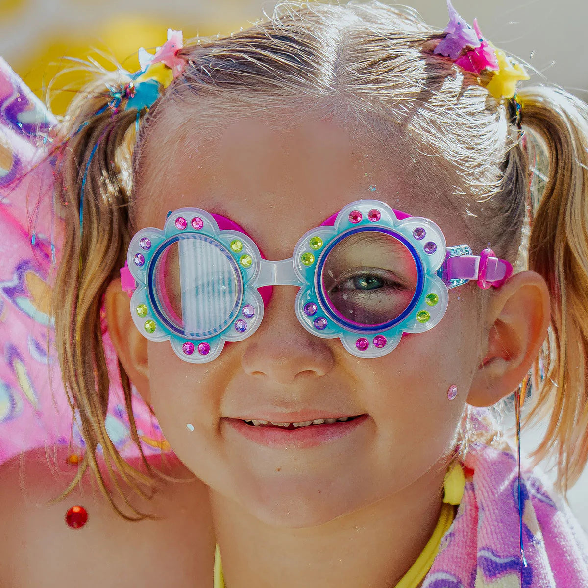Smiling at the camera, a child with blonde pigtails wears Bling2O Petals Swim Goggles featuring a jeweled, flower-shaped frame and has a pink towel draped around their shoulders.