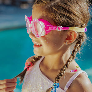 A young girl with braided hair and colorful beads wears Bling2O Petals Swim Goggles, featuring a flower-shaped frame, and a white swimsuit as she stands by a swimming pool.