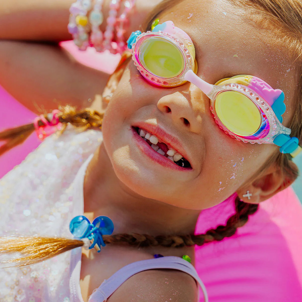 A young girl with braided hair smiles, showing missing front teeth, as she enjoys the sunshine outdoors wearing Bling2O Rainbow Swim Goggles with a glittery frame, along with bracelets and a necklace.