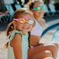 Two young girls in swimsuits, wearing Bling2O Kids' Crown Swim Goggles with vibrant UV protection, sit by the edge of a pool and smile at the camera on a sunny day.