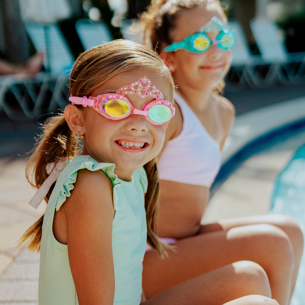 Two young girls in swimsuits, wearing Bling2O Kids' Crown Swim Goggles with vibrant UV protection, sit by the edge of a pool and smile at the camera on a sunny day.