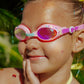 A young child wearing Bling2O's Tropical Rhinestone Swim Goggles smiles and touches their chin, with sunshine and greenery in the background.