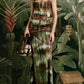 A woman stands before a tropical jungle backdrop wearing the Johanna Ortiz Pine Green Plant of Joy Ankle Dress and holding a brown handbag.