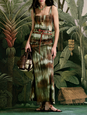 A woman stands before a tropical jungle backdrop wearing the Johanna Ortiz Pine Green Plant of Joy Ankle Dress and holding a brown handbag.