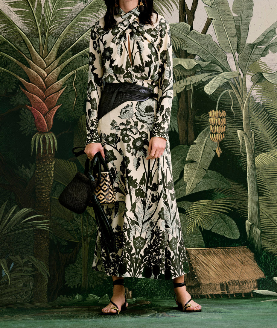 A woman wearing the Johanna Ortiz Heavenly Garden Top stands in front of a jungle-themed backdrop, holding a patterned handbag.