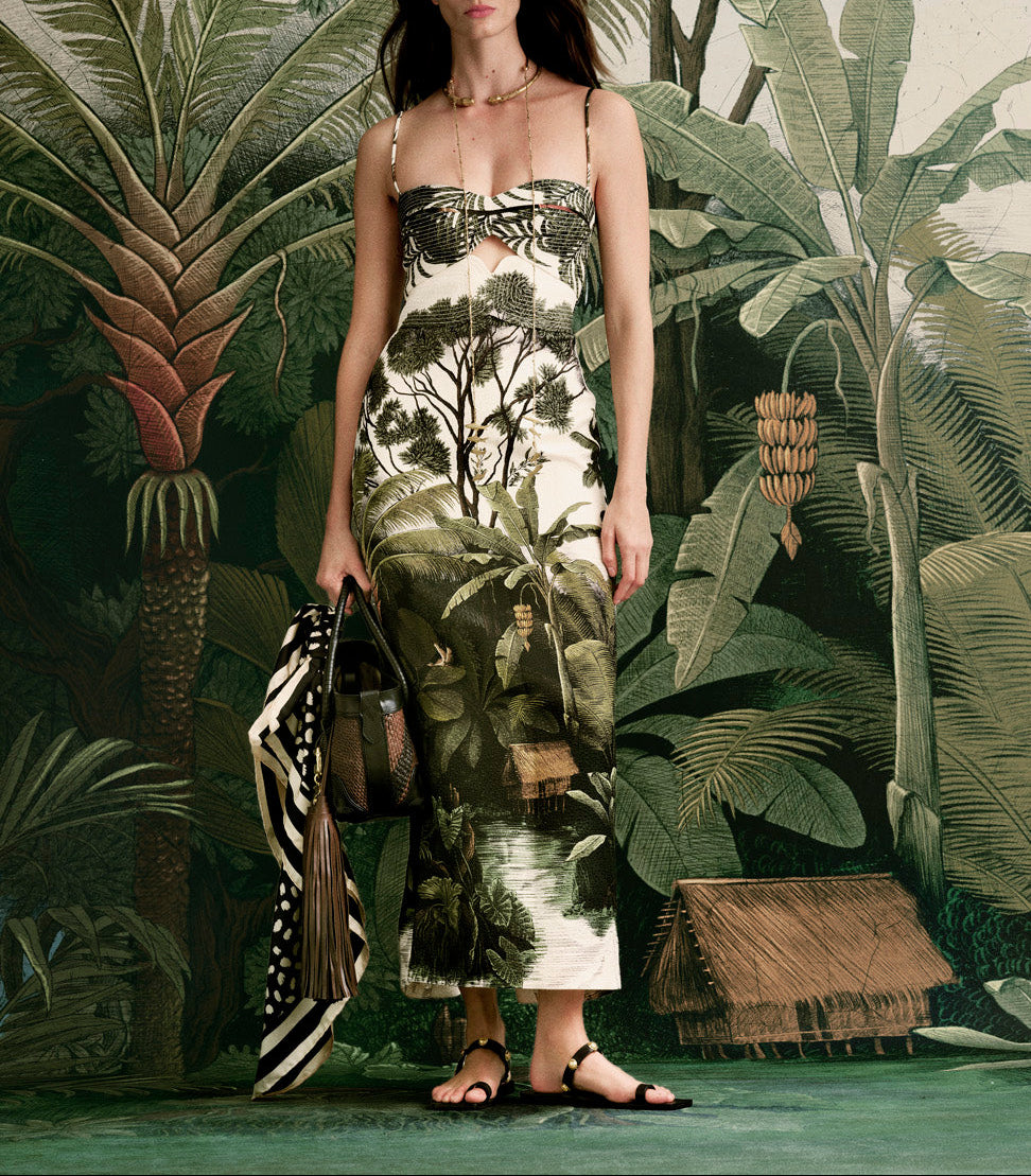 A woman in the Johanna Ortiz Tropicana Ankle Dress, featuring a Colombian landscape motif, stands before a jungle-inspired backdrop holding a patterned bag and scarf. Her face is not shown.
