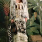 A woman in the Johanna Ortiz Invictus Ankle Dress stands before a jungle-themed backdrop, holding a handbag and scarf. The linen dress features botanical prints, layered ruffles, and an effortlessly chic style.