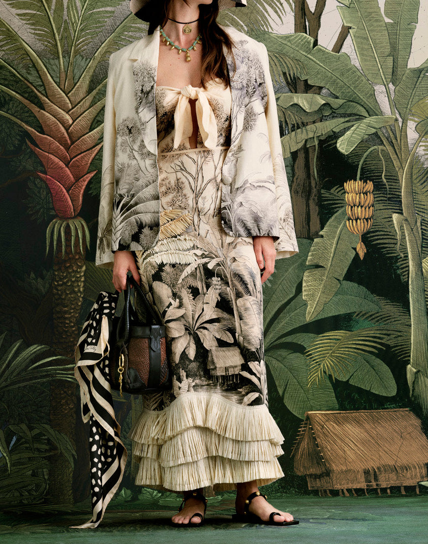 A woman in the Johanna Ortiz Invictus Ankle Dress stands before a jungle-themed backdrop, holding a handbag and scarf. The linen dress features botanical prints, layered ruffles, and an effortlessly chic style.