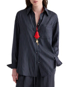 A person wearing the TWP Big Joe Evening Stripe Silk Shirt—an elegant, dark, menswear-inspired pinstriped button-up—paired with matching pants and accessorized with a red lanyard and key around their neck.