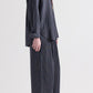 A person stands sideways against a plain light background, wearing the TWP Big Joe Evening Stripe Silk Shirt in a loose, menswear-inspired fit, paired with matching wide-leg pants and open-toe sandals.