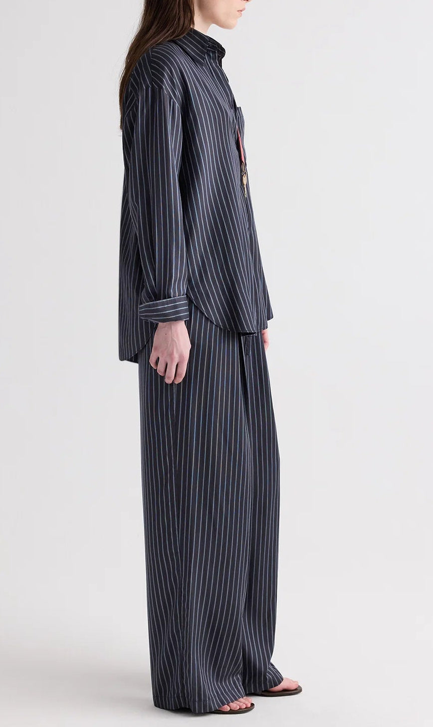 A person stands sideways against a plain light background, wearing the TWP Big Joe Evening Stripe Silk Shirt in a loose, menswear-inspired fit, paired with matching wide-leg pants and open-toe sandals.