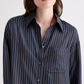 A person wearing the TWP Big Joe Evening Stripe Silk Shirt—a menswear-inspired black button-up featuring blue vertical stripes and a front chest pocket—shown from shoulders to mid-torso.