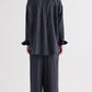 A person stands facing away, wearing the TWP Big Joe Evening Stripe Silk Shirt—a menswear-inspired, loose-fitting dark pinstripe button-down—paired with matching wide-leg pants. The relaxed silhouette of the shirt adds to the effortlessly chic look against the plain, light-colored background.
