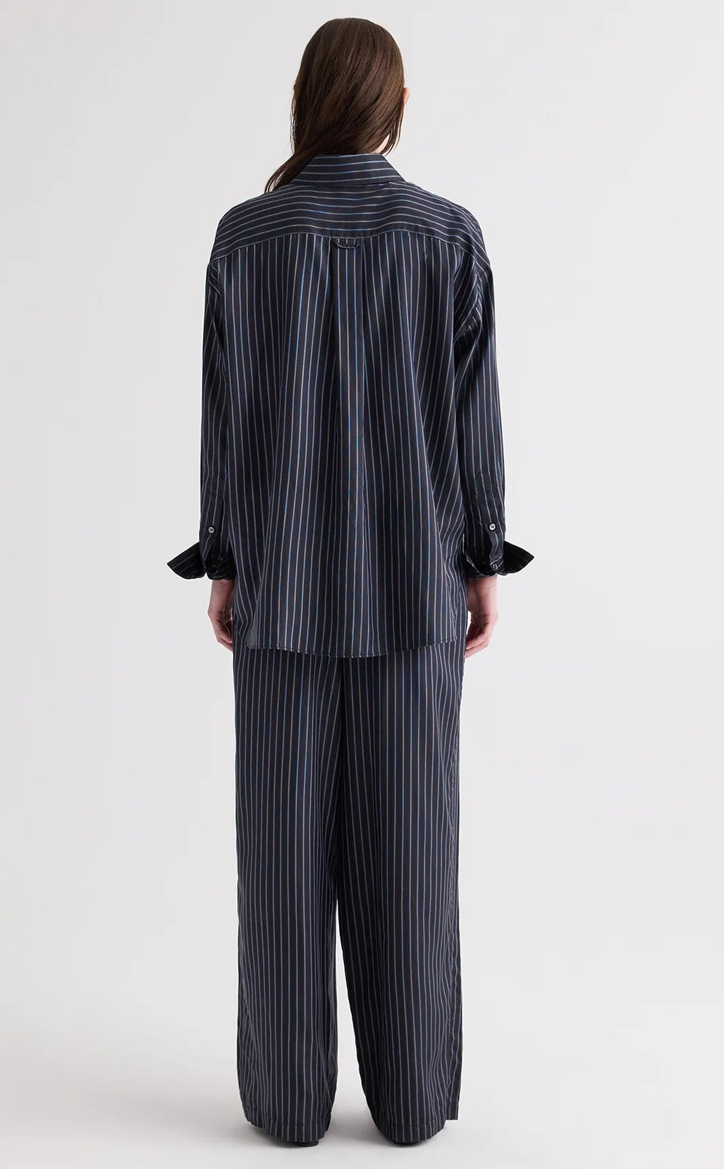 A person stands facing away, wearing the TWP Big Joe Evening Stripe Silk Shirt—a menswear-inspired, loose-fitting dark pinstripe button-down—paired with matching wide-leg pants. The relaxed silhouette of the shirt adds to the effortlessly chic look against the plain, light-colored background.