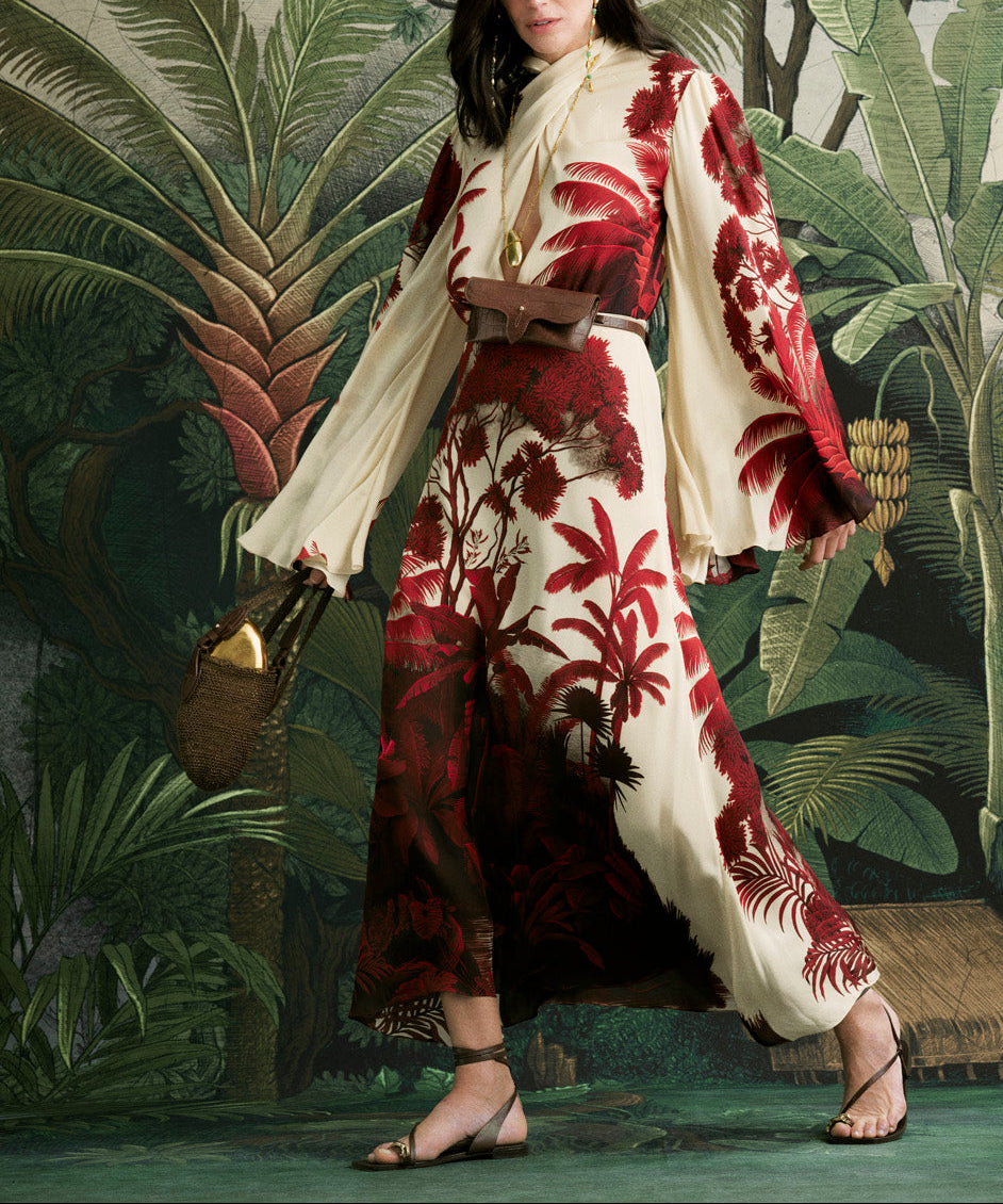 Wearing the Johanna Ortiz Maiden Voyage Ankle Dress, a woman stands before a tropical backdrop, accessorized with a brown belt bag and a woven clutch.