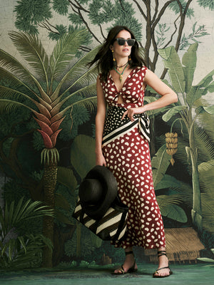 A woman in sunglasses wears the Johanna Ortiz Horizonte Mistico Ankle Dress with a red and white pattern, standing before a tropical mural while holding a large hat and striped bag.