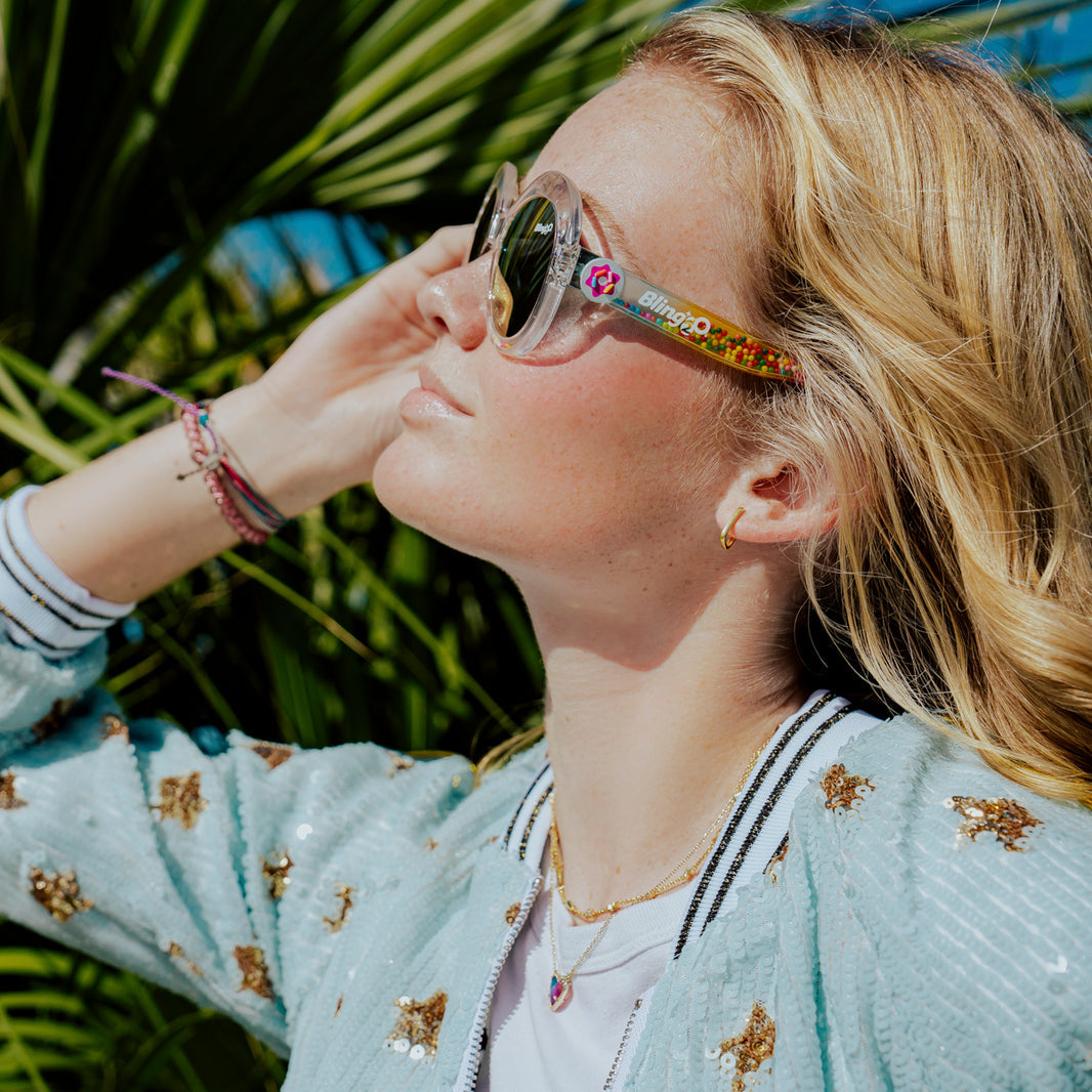 A woman stands outdoors in sunlight among green palm leaves, wearing Bling2O Girls’ Glass Beach Sprinkle Sunrise Sunglasses with UV-protected lenses and a light blue sequin jacket.