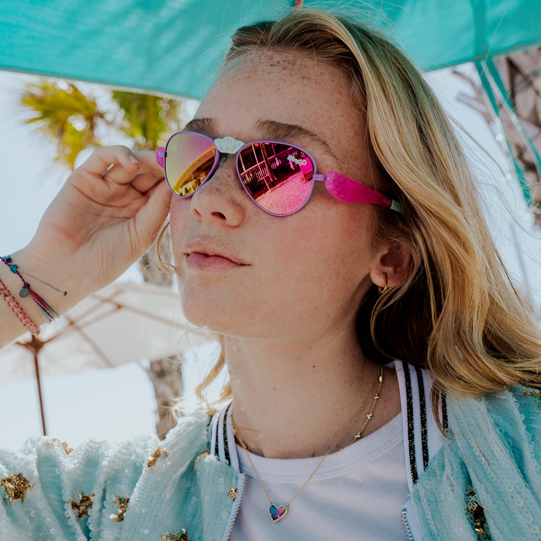 A young woman with long blonde hair wears Bling2O Girls’ Hampton Beach Sunglasses—stylish, reflective pink shades featuring UV-protected lenses—paired with a light blue jacket under a teal umbrella, as palm trees and umbrellas fill the sunny outdoor background.