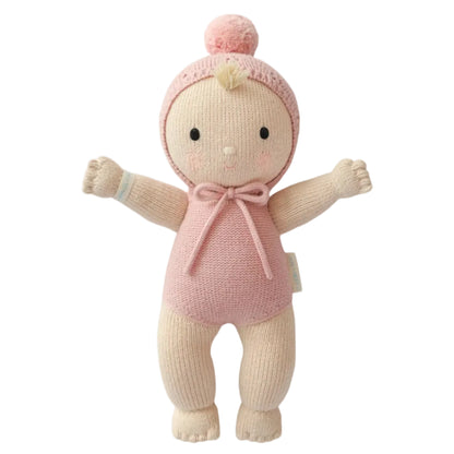 My First Baby by Cuddle + Kind is a stuffed baby doll featuring beige arms and legs crafted from natural cotton, dressed in a pink hand-knit outfit with a hood and pom-pom, standing upright with arms outstretched.