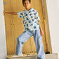 A boy wears Gingersnaps Boys' Loose Cargo Pant Denim with Banana Print and a white patterned shirt, standing on a doorstep and leaning against a wooden door.