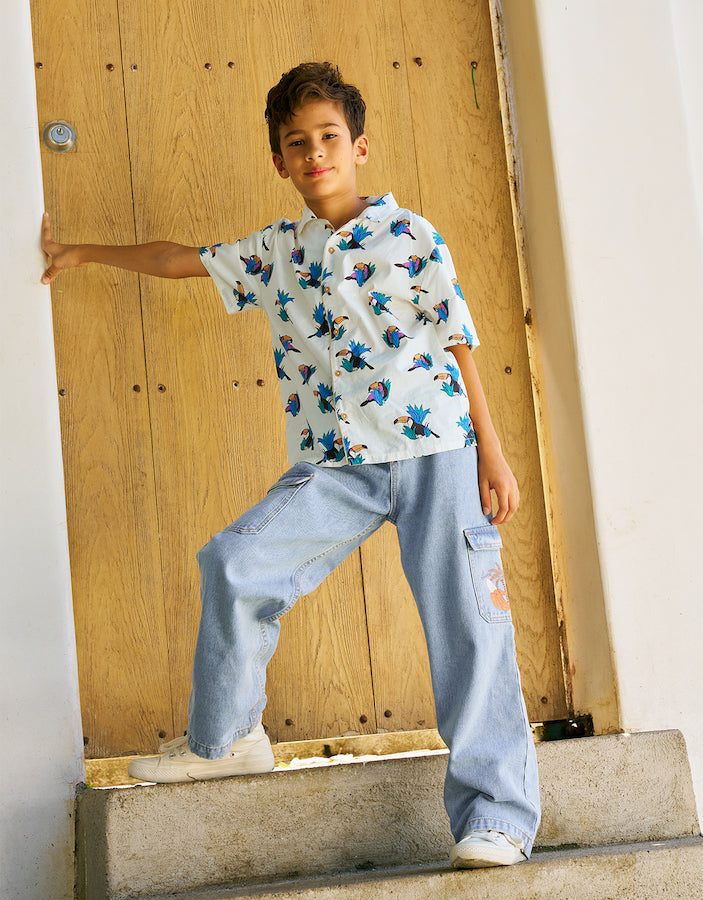 A boy wears Gingersnaps Boys' Loose Cargo Pant Denim with Banana Print and a white patterned shirt, standing on a doorstep and leaning against a wooden door.