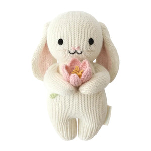 The Cuddle + Kind Baby Bunny - Blooming Tulip is an heirloom-quality, handcrafted cotton yarn plush bunny featuring black eyes, a small embroidered smile, and holding a pink tulip flower, displayed on a white background.