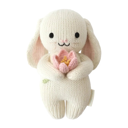 The Cuddle + Kind Baby Bunny - Blooming Tulip is an heirloom-quality, handcrafted cotton yarn plush bunny featuring black eyes, a small embroidered smile, and holding a pink tulip flower, displayed on a white background.