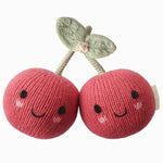 Two smiling, pink, knit cherry plush toys called “Baby Cherries” from Cuddle + Kind, featuring green knit leaves and joined by their stems, are handcrafted from cotton yarn and displayed on a plain white background.