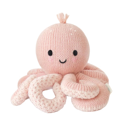 The Baby Octopus - Soft Coral Pink by Cuddle + Kind is a handcrafted stuffed toy made from soft Peruvian cotton yarn. Featuring a round head, black eyes, a smiling mouth, and eight curled tentacles, this adorable heirloom-quality animal is displayed against a white background.