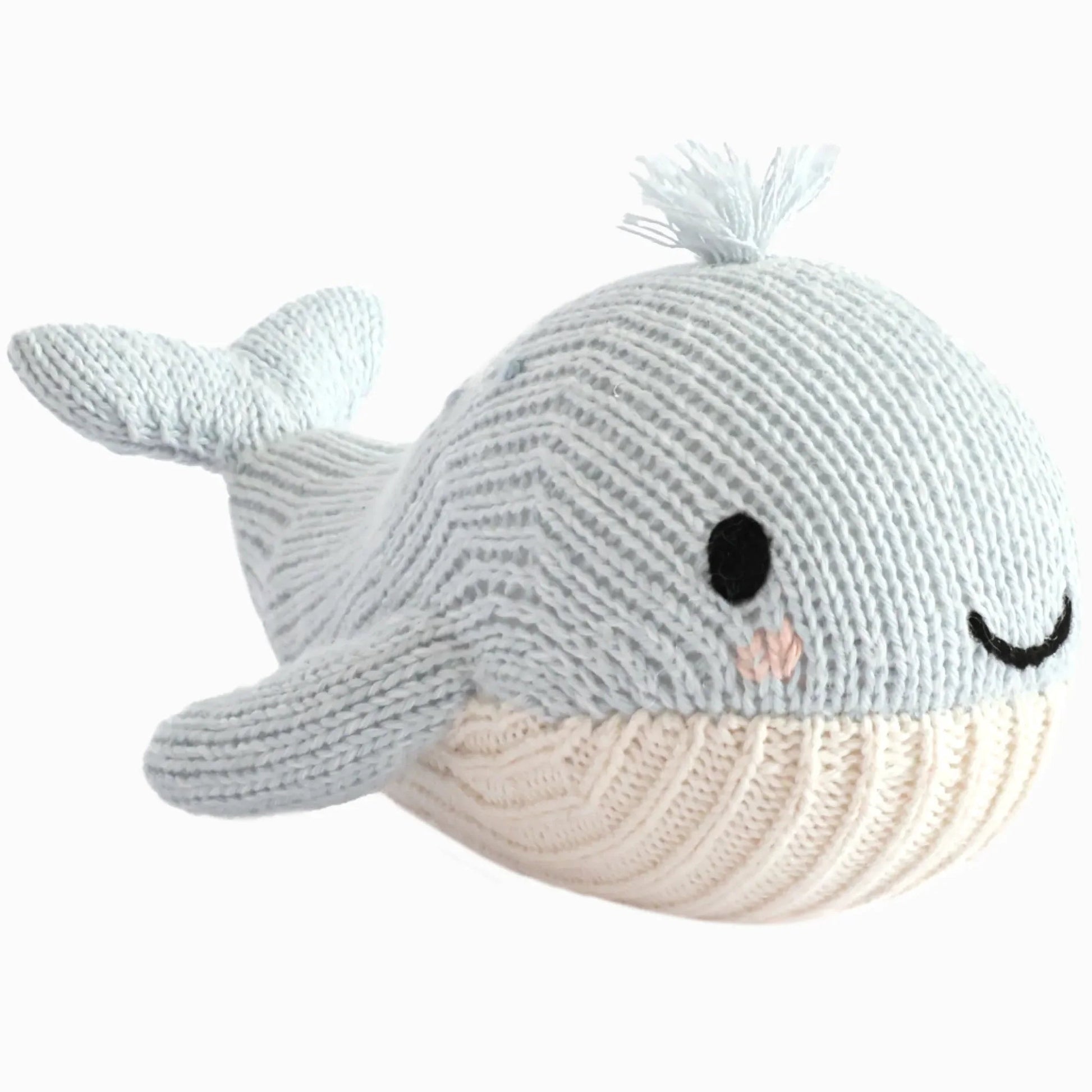 The Cuddle + Kind Baby Whale is a light blue knitted plush toy, handcrafted from heirloom-quality Peruvian cotton, featuring a white underside, black eyes, a pink blush spot, a smiling mouth, and a small knit tuft on its head—making it an irresistibly soft companion for babies.