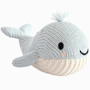 The Cuddle + Kind Baby Whale is a light blue knitted plush toy, handcrafted from heirloom-quality Peruvian cotton, featuring a white underside, black eyes, a pink blush spot, a smiling mouth, and a small knit tuft on its head—making it an irresistibly soft companion for babies.