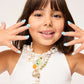 A young girl in a white top shows off her ocean-inspired blue “Mermaid Press On Nail Kit” by Super Smalls and wears layered pearl necklaces with sea-themed pendants, smiling at the camera.