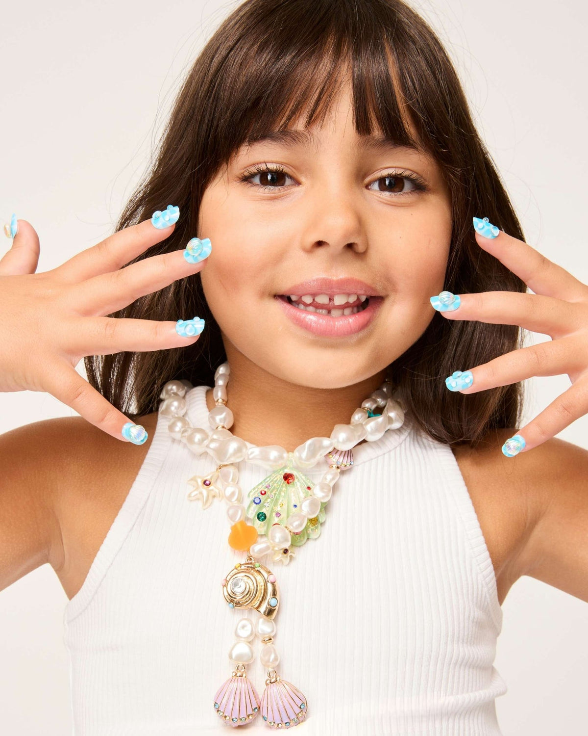 A young girl in a white top shows off her ocean-inspired blue “Mermaid Press On Nail Kit” by Super Smalls and wears layered pearl necklaces with sea-themed pendants, smiling at the camera.