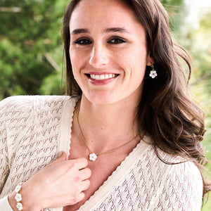 A woman outdoors wears a cream knit sweater, gold Juliska Capucine De Wulf Mermaid Garden Hoop Earrings with Flower Charm, a gold flower necklace, and a matching bracelet, with greenery in the background.