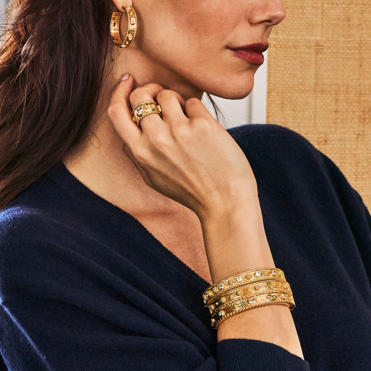 A woman in a dark blue top wears gold hoop earrings, a thick gold ring, and two Juliska Capucine De Wulf Berry Classic Hinged Bangles stacked as bracelets with embedded stones.