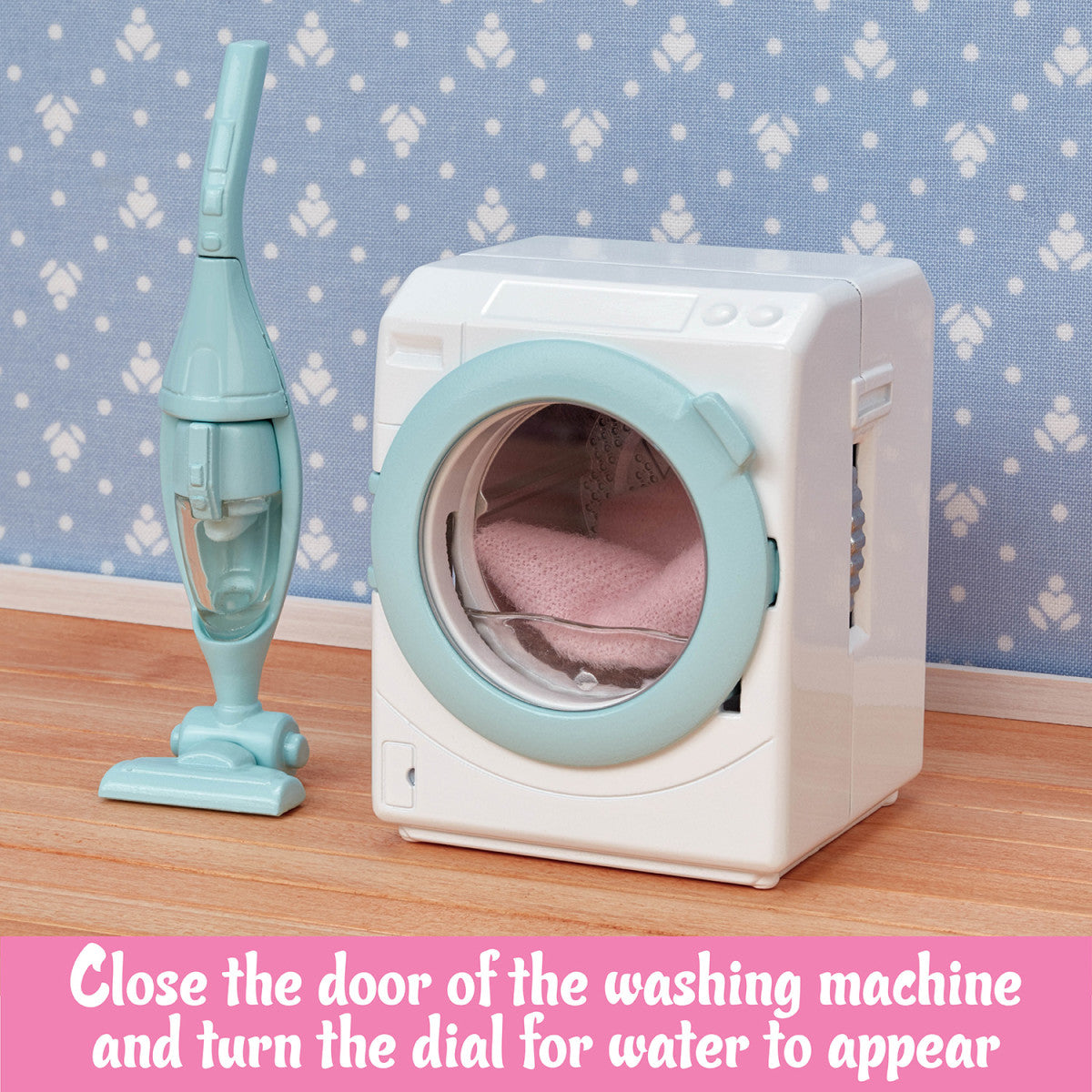 The Calico Critters Laundry & Vacuum Cleaner set features a toy washing machine with a pink towel inside, placed beside a small blue toy vacuum on a wooden floor against a blue patterned wall—making it the perfect charming accessory set for your dollhouse collection.